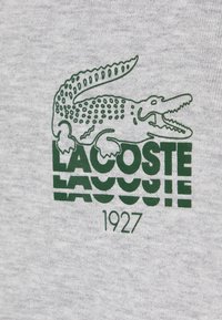 Tessuto di cotone grigio con logo del coccodrillo verde Lacoste e testo del marchio, con l'anno "1927" stampato sotto in caratteri in grassetto.