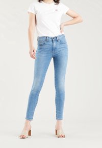 Pantalones vaqueros ajustados de mezclilla azul con cintura alta, diseño de cinco bolsillos y un ligero desgaste. Combinados con una camiseta blanca ajustada y sandalias beige de tacón bloque.