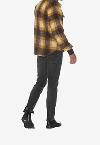 Gul og brun ternet flannel skjorte, sorte jeans og sorte støvler. Skjorten har en afslappet pasform med knaplukning og synlige strukturer.