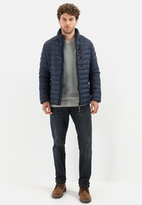 Marineblauw gewatteerd pufferjack met een hoge kraag, een grijze trui eronder, donkere jeans en bruine laarzen. Soepele textuur met een ritssluiting aan de voorkant.