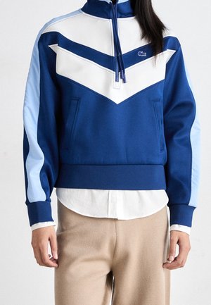 Kvinde iført en blå og hvid kvart lynlås trøje med Chevron-design, beige bukser og hvid skjorte under.