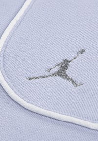 Jasnofioletowa tkanina o fakturowanej powierzchni; z wyraźnym srebrnym haftowanym logo Jordana i białym wykończeniem wzdłuż krawędzi.