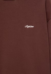 Gros plan sur un tissu marron avec un petit texte blanc brodé « "Righttime" » au niveau de la poitrine d'un vêtement.