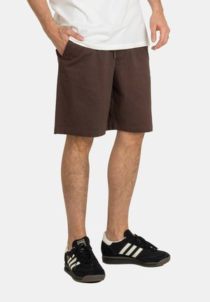 Mann trägt braune knielange Shorts, weißes Hemd und schwarze Adidas-Sneaker mit weißen Streifen, steht vor einem einfarbigen Hintergrund.