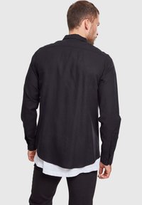 Camisa negra de manga larga con cuello estructurado, puños abotonados y un corte relajado, usada sobre una camisa blanca para un efecto de capas.