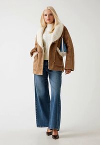 Bruine shearlingjas met een brede witte kraag, gecombineerd met een crème kabelgebreide trui en een blauwe hoge jeans. Bruine hakken.