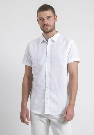 Homme portant une chemise blanche à manches courtes et un pantalon blanc, debout devant un fond gris clair uni.