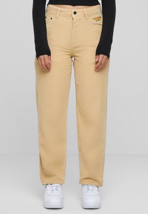 Beige Cord-Jeans mit weitem Bein, frontalen Taschen, Reißverschluss und gestickten Details. Kombiniert mit weißen Sneakers.