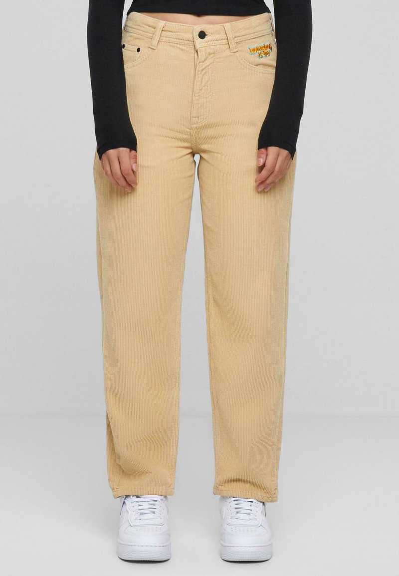 Pantaloni in corduroy beige a gamba larga con tasche anteriori, zip e dettagli ricamati. Abbinati a sneakers bianche.