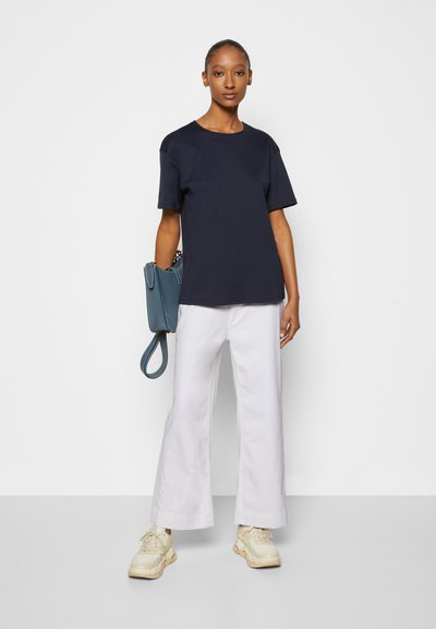 T-shirt de gola redonda azul-marinho, mangas curtas, combinado com calças largas brancas e ténis bege; segurando uma bolsa azul-petróleo. Design simples e moderno.