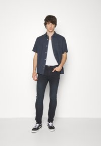Camisa de botones de manga corta en color marino sobre una camiseta blanca, combinada con pantalones vaqueros ajustados oscuros y zapatillas negras con acentos blancos.