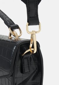 Sac à main en cuir noir texturé avec fermoir en métal doré fixant la bandoulière au corps du sac.