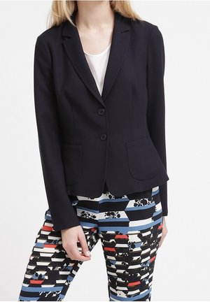 Femme portant un blazer noir ajusté sur un haut blanc avec un pantalon à rayures multicolores et motifs floraux, posant devant un fond blanc.