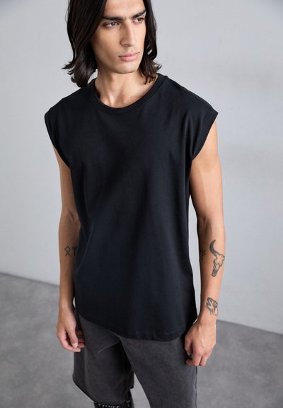 Zwarte mouwloze T-shirt van zacht, lichtgewicht katoen. Beschikt over een ronde halslijn en een relaxte pasvorm, met zichtbare tatoeages op de arm.
