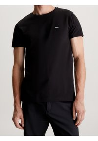Musta puuvillainen t-paita, jossa on lyhyet hihat, pyöreä pääntie ja pieni logoplaatta vasemmassa rinnassa. Sileä pinta ja vartalonmyötäinen malli.