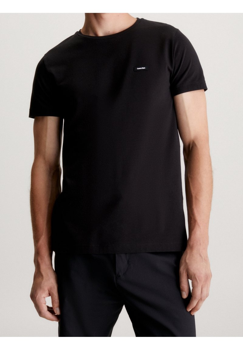 Musta puuvillainen t-paita, jossa on lyhyet hihat, pyöreä pääntie ja pieni logoplaatta vasemmassa rinnassa. Sileä pinta ja vartalonmyötäinen malli.