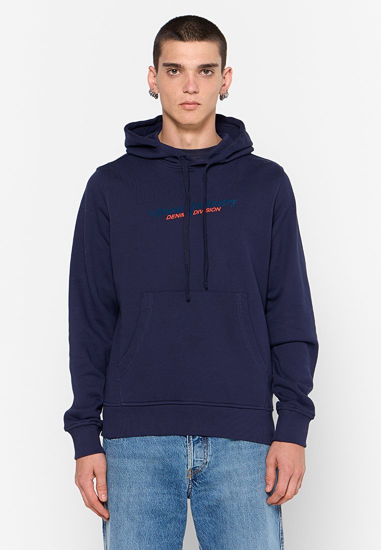 Jeune homme portant un sweat à capuche bleu marine avec poche avant et un jean bleu, se tenant contre un fond blanc uni.