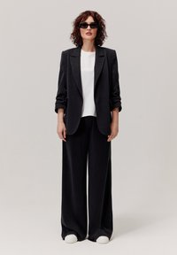 Czarna marynarka idealnie dopasowana na białym t-shircie, zestawiona z szerokimi czarnymi spodniami i białymi sneakersami. Model nosi ciemne okulary przeciwsłoneczne.