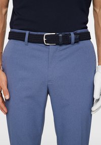 Marineblauer gewebter Gürtel mit silberner Metallschnalle, getragen mit hellblauen, strukturierten Hosen. Eine Hand in einem weißen Handschuh ruht auf dem Oberschenkel.