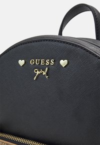 Musta tekstuuriga seljakoti osa, kus on kullast "GUESS girl" logo ja kaks väikest kullast südamekujulist nupukest kullase erineva pehme luku kohal.