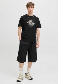 Černé tričko s krátkým rukávem a logem "JACK & JONES", doplněné černými kraťasy do kolen a béžovými teniskami, které se skvěle hodí k bílým vzorovaným ponožkám.