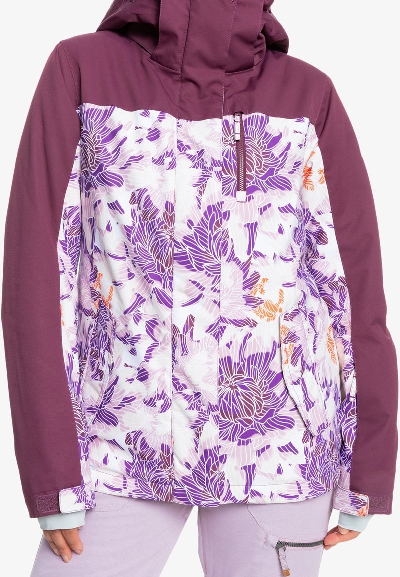 Lila och vit blommig jacka med enfärgade lila ärmar och bröst, framtida dragkedjeficka, bärs med ljuslila byxor med en dragkedjeficka.