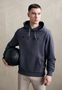 Hoodie en coton gris avec une poche kangourou, capuche à cordon de serrage et logo "Champion" brodé. Le modèle tient une balle noire ronde.