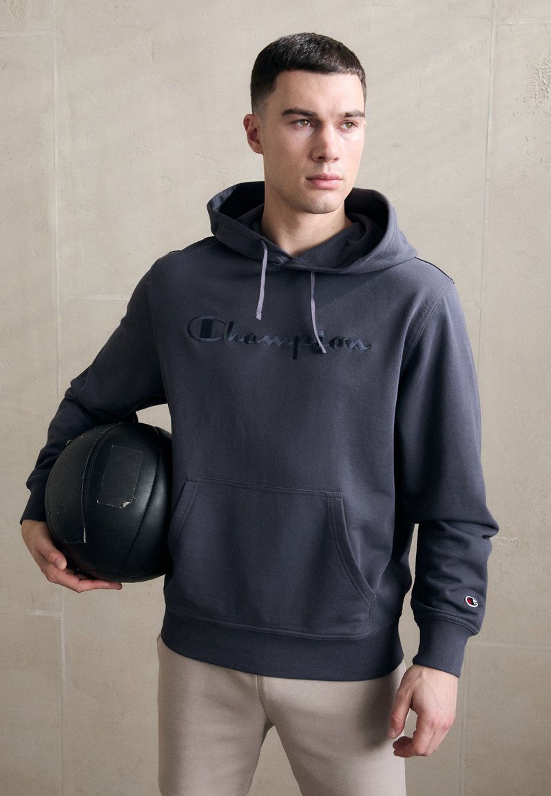 Hoodie en coton gris avec une poche kangourou, capuche à cordon de serrage et logo "Champion" brodé. Le modèle tient une balle noire ronde.