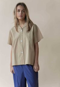 Beige Kurzarmhemd mit Reverskragen und Knöpfen, kombiniert mit blauen Hosen mit Pünktchenmuster, die weite Beine und eine entspannte Passform haben.