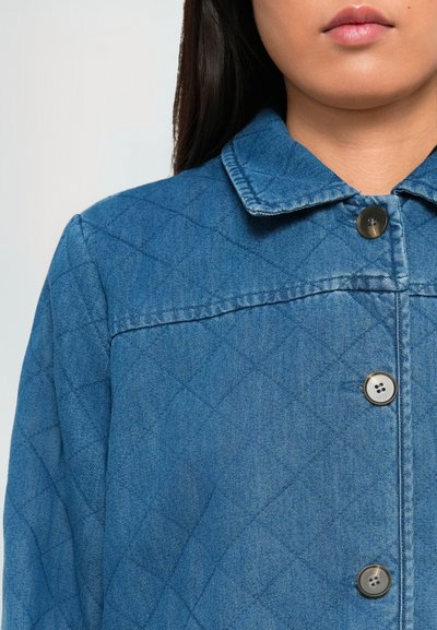Femeie îmbrăcată cu o jachetă bleu din denim matlasată, cu butoane mari, vizibilă de la gât până la jumătatea toracelui, pe un fundal simplu.