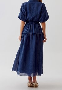 Maxiklänning i marinblått, tillverkad av texturerat tyg. Har en figurnära midja, puffärmar och en lager-på-lager fåll. Bärs med ljusa högklackade skor.