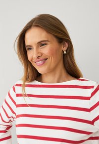 Camisa blanca de manga larga con rayas horizontales rojas. El modelo tiene el cabello castaño ondulado y lleva pendientes de oro. Fondo neutro.
