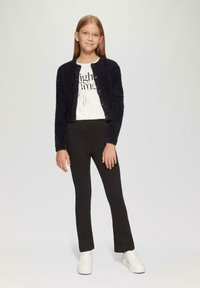 Schwarzer, flauschiger Cardigan mit Knöpfen, weißes T-Shirt mit Grafik, schwarze Leggings und weiße Sneaker. Model steht mit den Händen an den Seiten.
