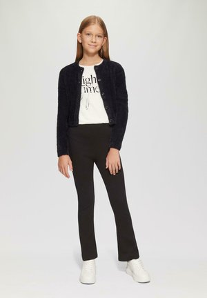 Czarny puszysty kardigan z guzikami, biały t-shirt z grafiką, czarne legginsy i białe sneakersy. Model stoi, ręce wzdłuż ciała.