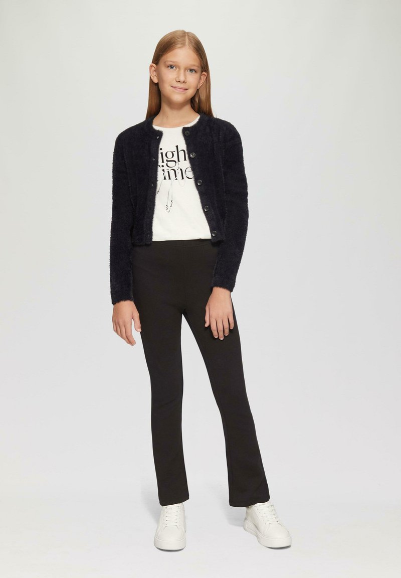 Schwarzer, flauschiger Cardigan mit Knöpfen, weißes T-Shirt mit Grafik, schwarze Leggings und weiße Sneaker. Model steht mit den Händen an den Seiten.