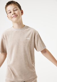 Camiseta de rizo beige con mangas cortas, cuello redondo y pequeño logo blanco en el pecho. Textura suave, corte relajado y diseño minimalista.