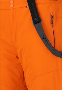 Pantalon imperméable orange avec une texture lisse, doté d'une poche zippée sur le côté, de deux fermetures boutonnées et d'une sangle noire réglable.