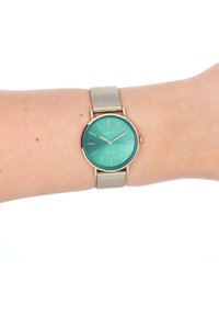 Montre à cadran teal avec des accents dorés, boîtier rond et bracelet en maille argentée. Dispose de marqueurs d'heure minimalistes et d'un logo de la marque.