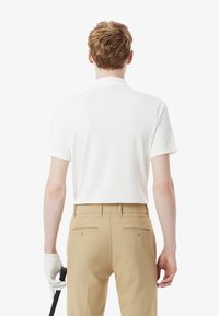 Polo blanc à manches courtes avec un col plat, associé à un pantalon beige doté de deux poches arrière, tenu par une main portant un gant blanc.