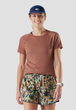 Uśmiechnięta kobieta w granatowej czapce, rdzawym krótkim rękawie, kolorowych kwiatuszkowych szortach i białym zegarku na nadgarstku, stojąca na jednolitym szarym tle.