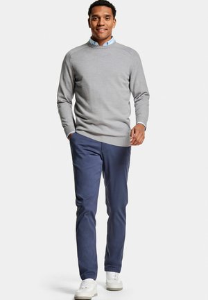 Man loopt zelfverzekerd, draagt een grijze trui over een lichtblauw hemd met kraag, marineblauwe broek en witte sneakers op een effen witte achtergrond.