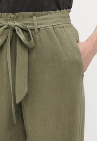 Olivově zelené, lehké kalhoty z texturované látky s elastickým pasem na šňůrku a bočními kapsami. Viditelný uzel na opaskové šňůrce.