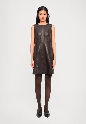 Woman wearing sleeveless dark brown leather dress, matching opaque tights, and black pointed-toe heels, standing against plain light background.