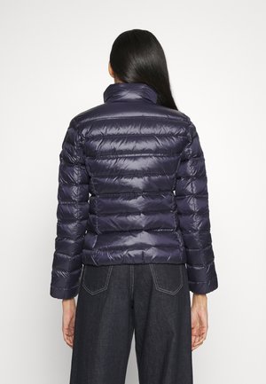 Femme aux longs cheveux foncés vue de dos, portant une doudoune bleu marine brillante et un pantalon en jean foncé sur un fond uni.