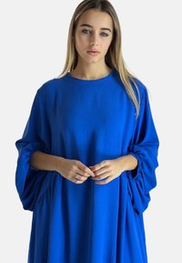 Blå oversized kjole med rund halsudskæring, tekstureret stof og voluminøse ærmer samlet ved manchetterne, hvilket skaber en flydende silhuet.