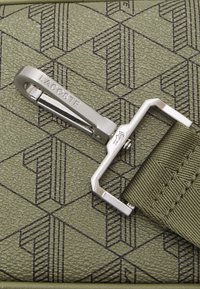Clip metallico con incisione "LACOSTE" su un cinturino verde oliva con un motivo testurizzato di forme geometriche.