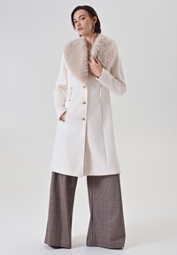 Cappotto bianco con colletto di pelliccia, bottoni dorati e tasche laterali. Abbinato a pantaloni a gamba larga a quadri con motivo houndstooth.