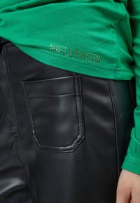 Grünes Baumwollhemd mit "INFLUENCER" in goldenen Strasssteinen. Kombiniert mit schwarzen Kunstlederhosen, die über eine Seitentasche und eine glatte Textur verfügen.