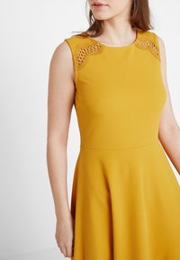 Žluté bezramínkové šaty s přiléhavým živůtkem a rozšířenou sukní. Pozoruhodné krajkové detaily na ramenou. Hladká, texturovaná tkanina.
