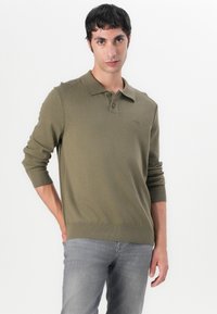 Olivgrüner Strickpullover mit Kragen, Knopfleiste mit drei Knöpfen, gerippten Bündchen und Saum. Verfügt über ein dezentes Logo-Detail auf der Brust.